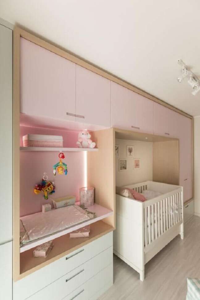Quarto de bebê com armário rosa apoiado na parede, com troçador embutido. Berço branco colocado sob o armário